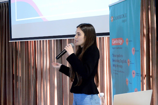 A woman in a black jacket speaking into a microphone on a stage at a conference.