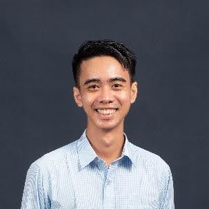 A young man with short black hair, wearing a light blue checkered shirt, smiling while posing in front of a plain dark background.