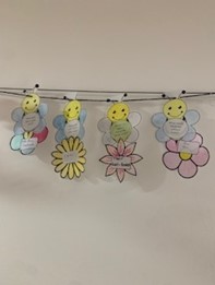 Four paper bee and flower crafts with handwritten notes are hanging on a string against a plain wall.