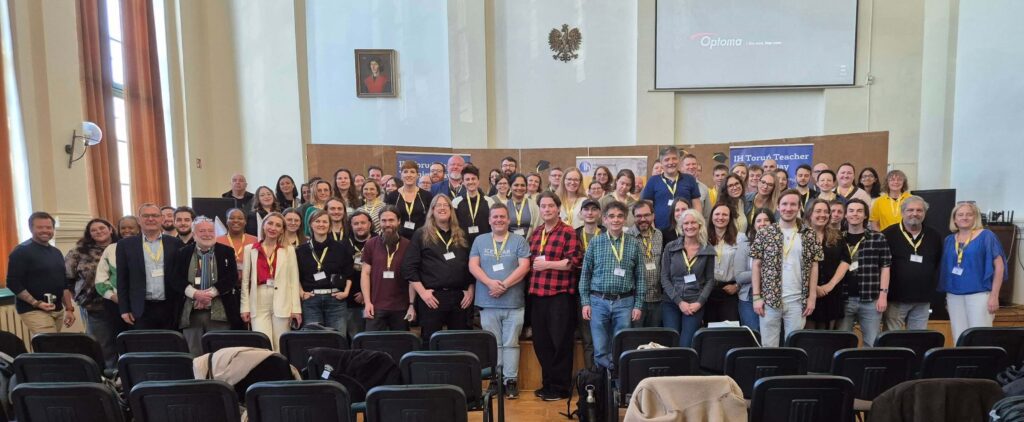 A large group of people stands posing for a group photo in a lecture hall with chairs, banners, and a projector screen in the background.