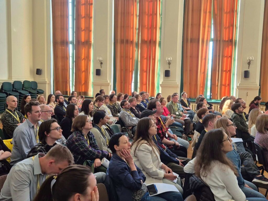 A group of people seated in rows, attentively listening to a presentation in a large room with tall windows and orange curtains.
