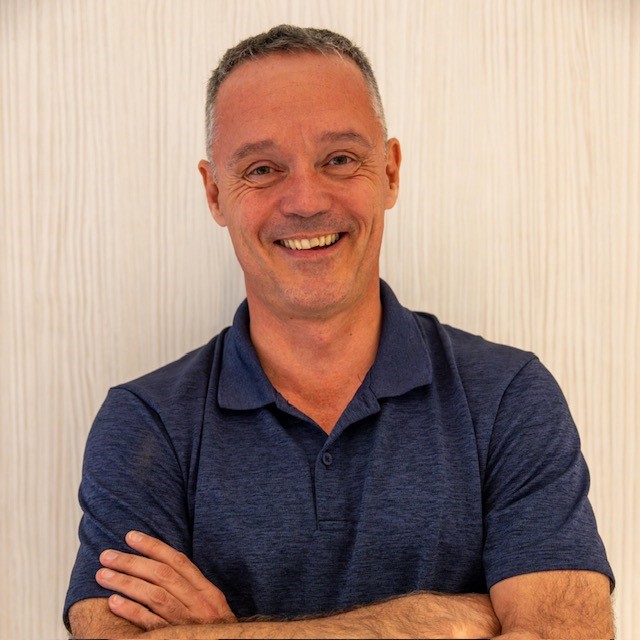 A man with short gray hair wearing a dark blue polo shirt stands smiling with his arms crossed against a light-colored, textured background.