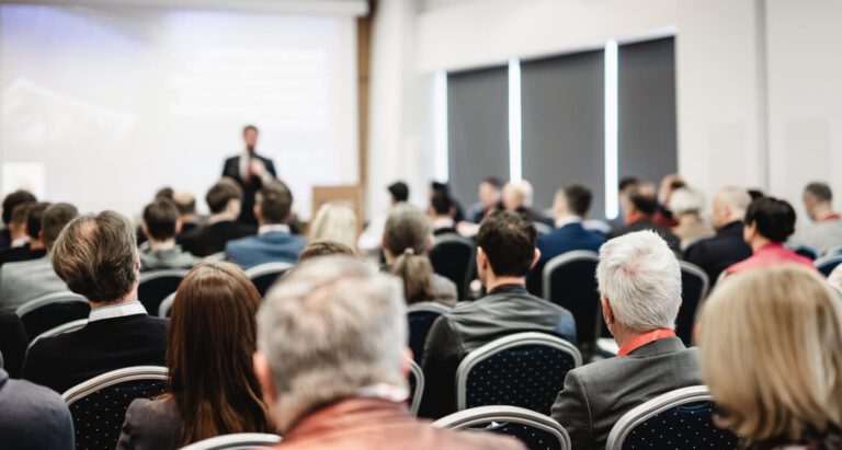 A group of people sit in rows of chairs facing a speaker giving a presentation in a conference room.