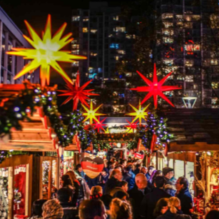 A crowded night scene at a European-style Christmas market, with wooden stalls, people mingling, and large, illuminated red and yellow star decorations hanging overhead.