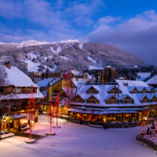 A snow-covered village square at dusk, possibly Whistler or a similar ski resort. Buildings are illuminated with festive red holiday lights, and snow-capped mountains rise in the background under a blue, cloudy sky.
