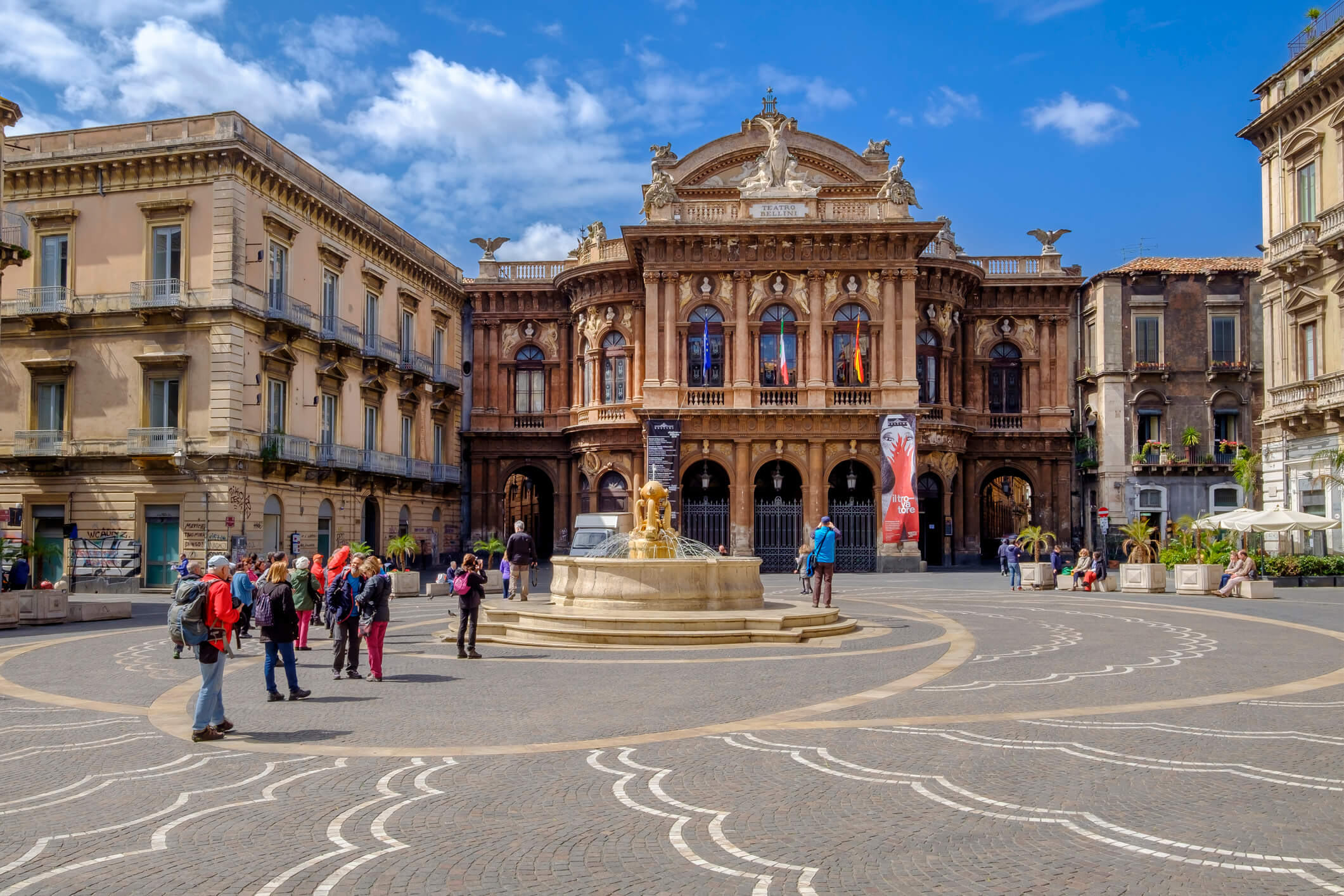 Baroque building with ornate facade facing a piazza with a circular fountain and people.