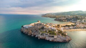 Aerial view of a coastal town with a historic stone fortress on a rocky peninsula, surrounded by the sea, beaches, and hills in the background.