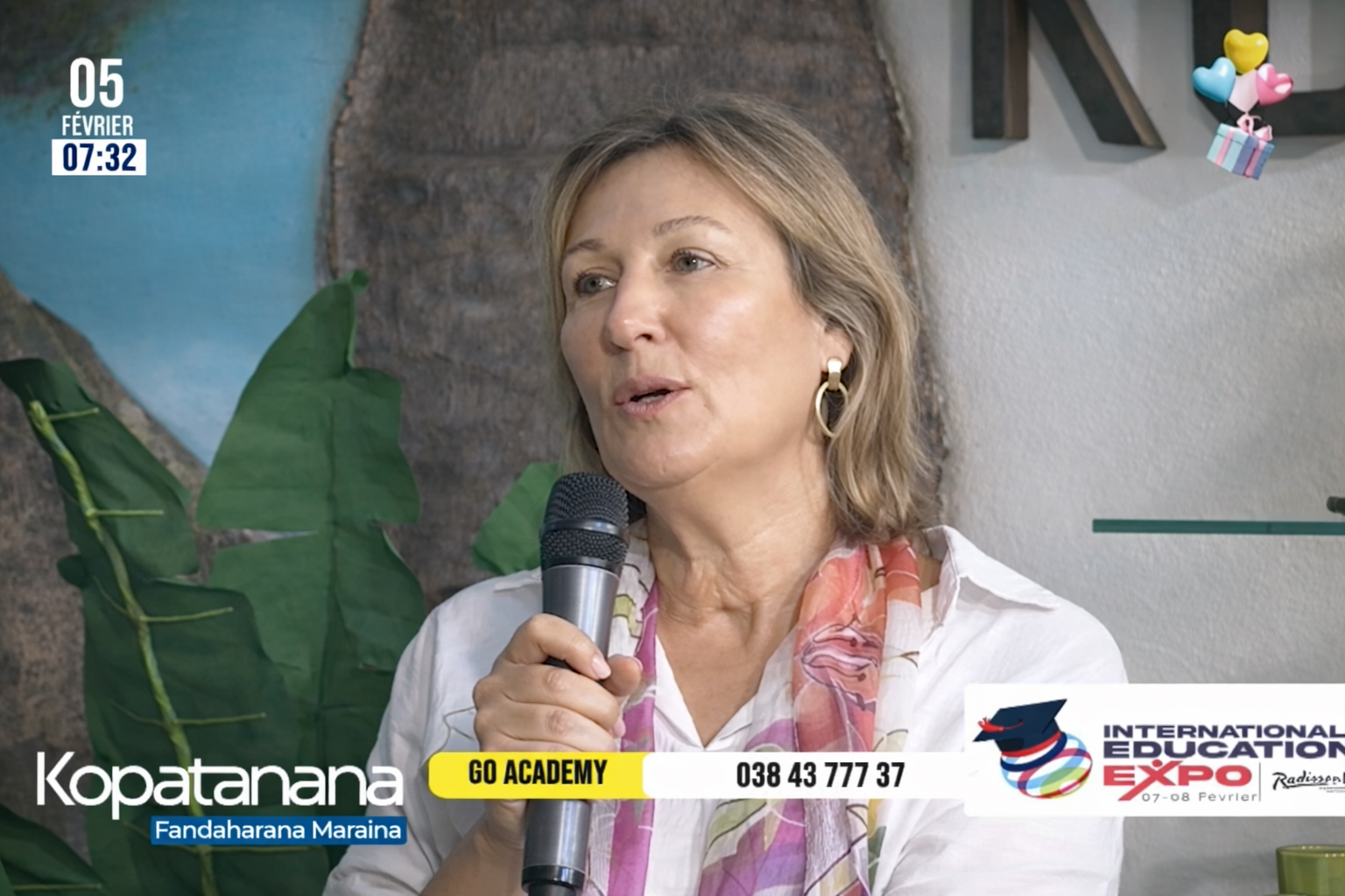 A woman with light hair and a colorful scarf speaks into a microphone during a TV segment. Event banners and contact info are displayed on the screen.