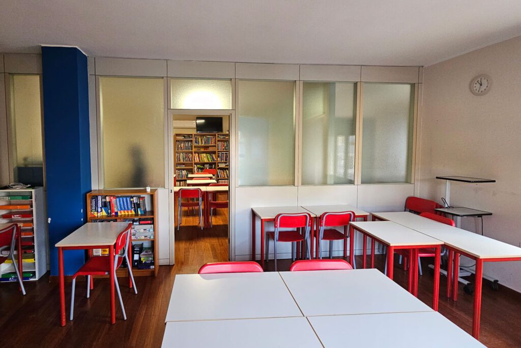 Classroom with white tables and red chairs, bookshelves along the left wall, and a view into a library through glass-paneled doors in the background.