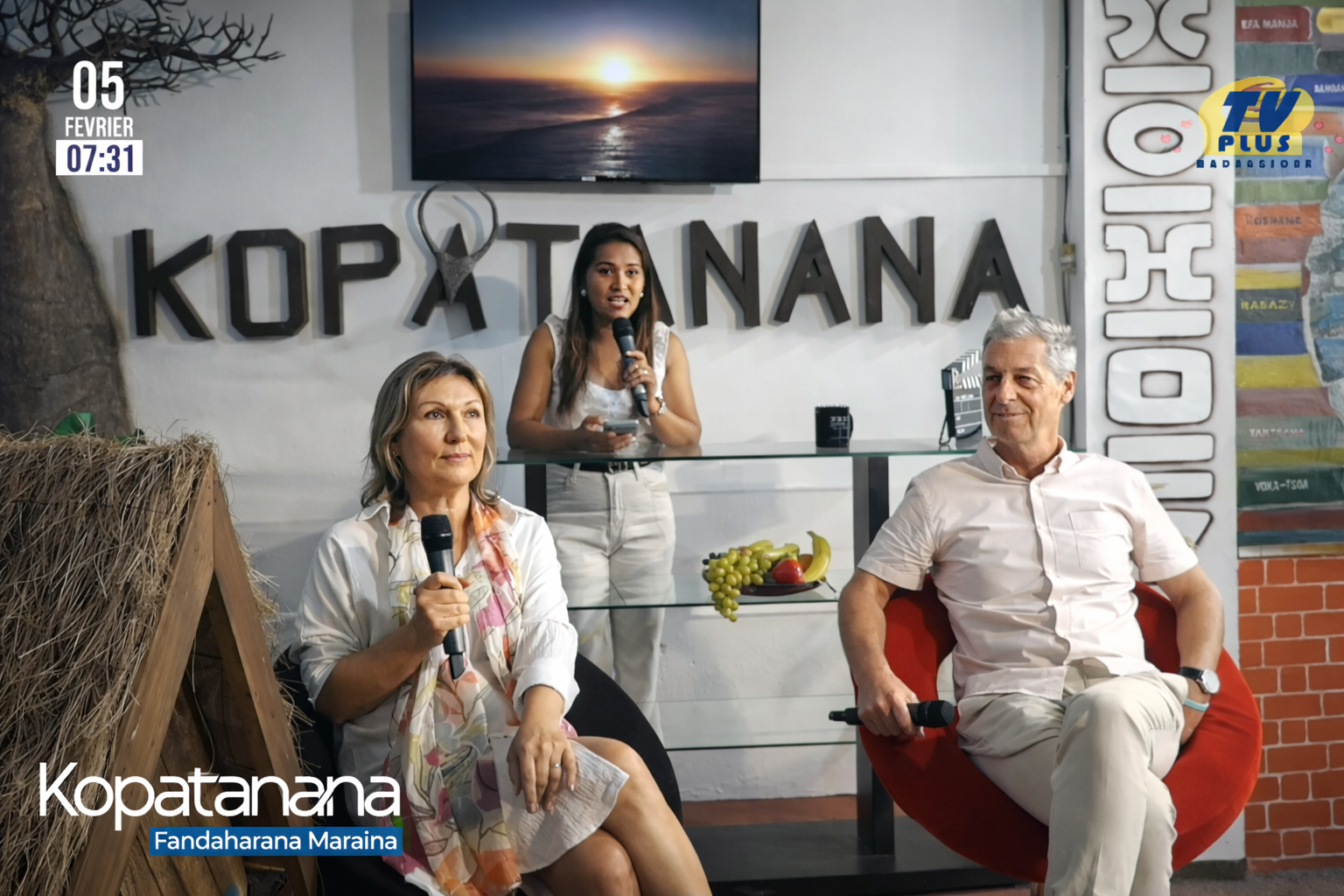 Three people participate in a TV interview set. Two sit with microphones in the foreground while a woman stands and speaks in the background. The text "Kopatanana" is displayed behind them.