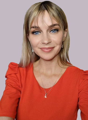 A woman with blonde hair and blue eyes is smiling, wearing a red top with puffed sleeves, drop earrings, and a necklace, against a plain light purple background.
