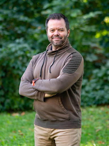A man with a beard stands outdoors on grass, wearing a brown sweater and tan pants, with arms crossed and greenery in the background.