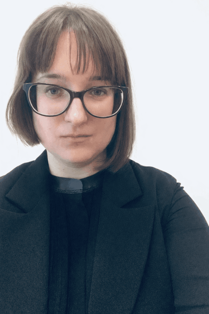 A person with straight brown hair, glasses, and a black jacket looks directly at the camera against a plain light background.
