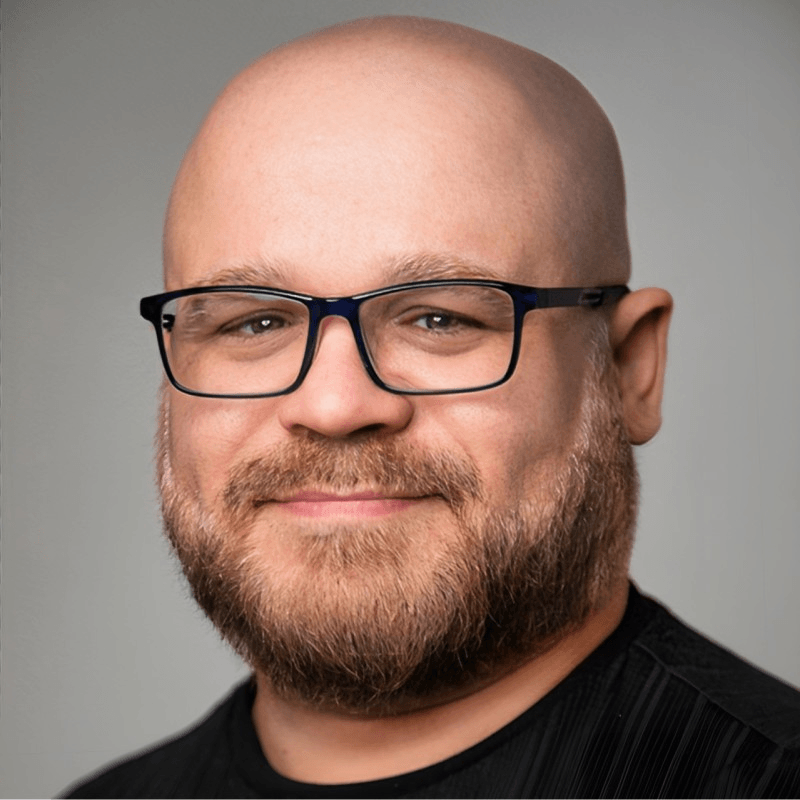 A man with a bald head, full beard, and glasses is smiling slightly while wearing a black shirt against a plain background.