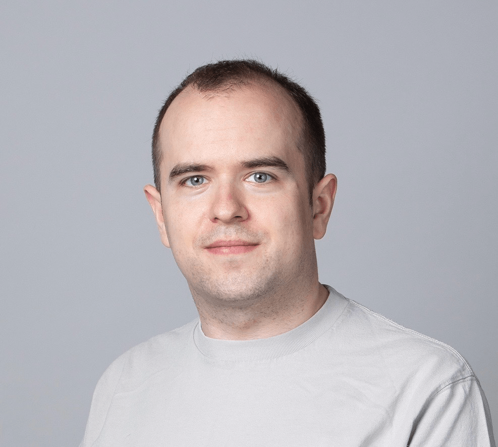 A man with short brown hair and light skin wearing a light gray t-shirt poses in front of a plain gray background.