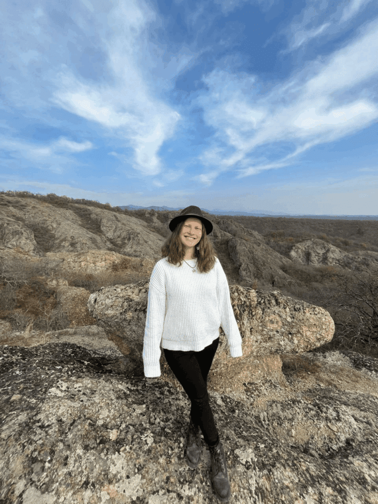A woman in a white sweater, black hat, and black pants stands on rocky terrain with hills and a blue sky in the background.