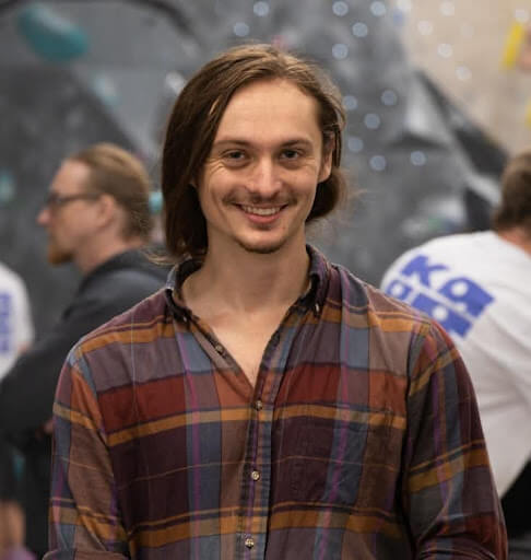 A man with shoulder-length brown hair and a plaid shirt smiles at the camera; there are people and a blurred background behind him.