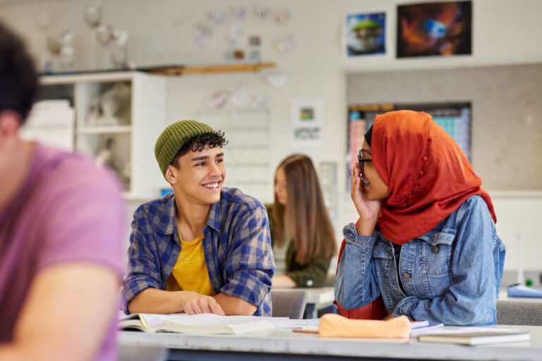 Two students sit at a classroom desk, smiling and talking. One wears a green beanie and plaid shirt; the other wears glasses and an orange hijab. Open books are on the desk.