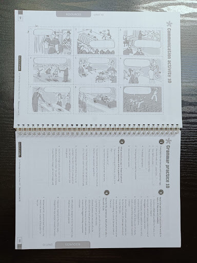 An open spiral-bound workbook on a dark table shows a comic strip activity on the top page and a grammar exercise on the bottom page.