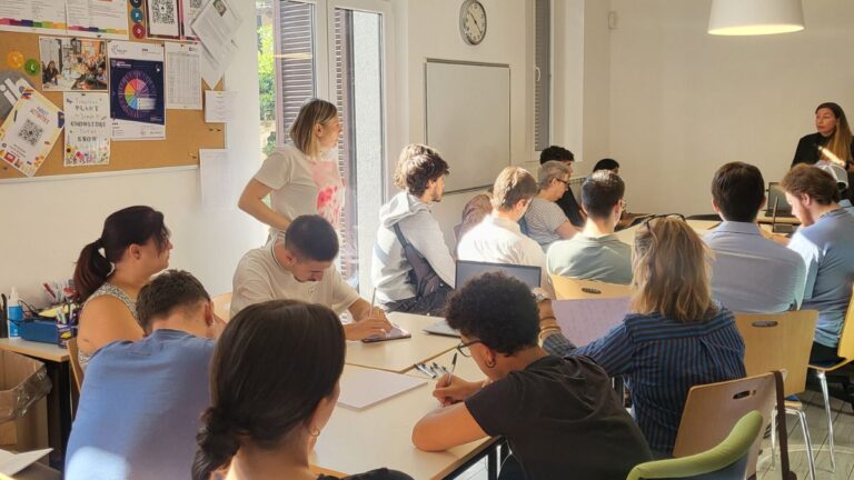 A group of people sit at desks in a well-lit classroom, listening to a woman speaking at the front near a whiteboard. Some attendees are taking notes.