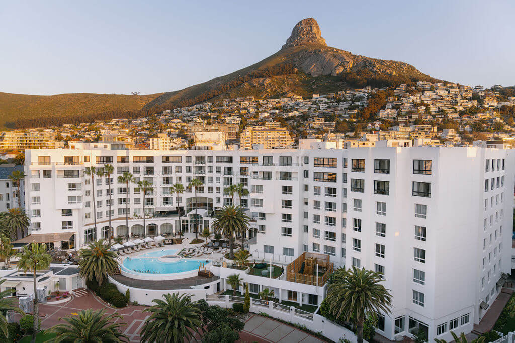 A large white hotel with a central pool, surrounded by palm trees, is set against a mountain and hillside dotted with houses under clear skies.
