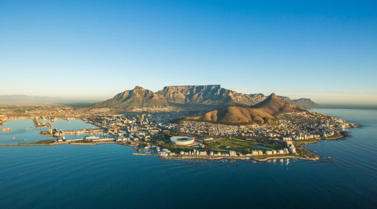 Aerial view of Cape Town with Table Mountain in the background, the cityscape, and coastline under clear blue skies.