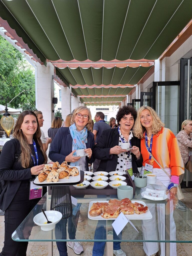 Four women stand around a glass table with pastries and snacks, smiling at an outdoor event under a green striped canopy.