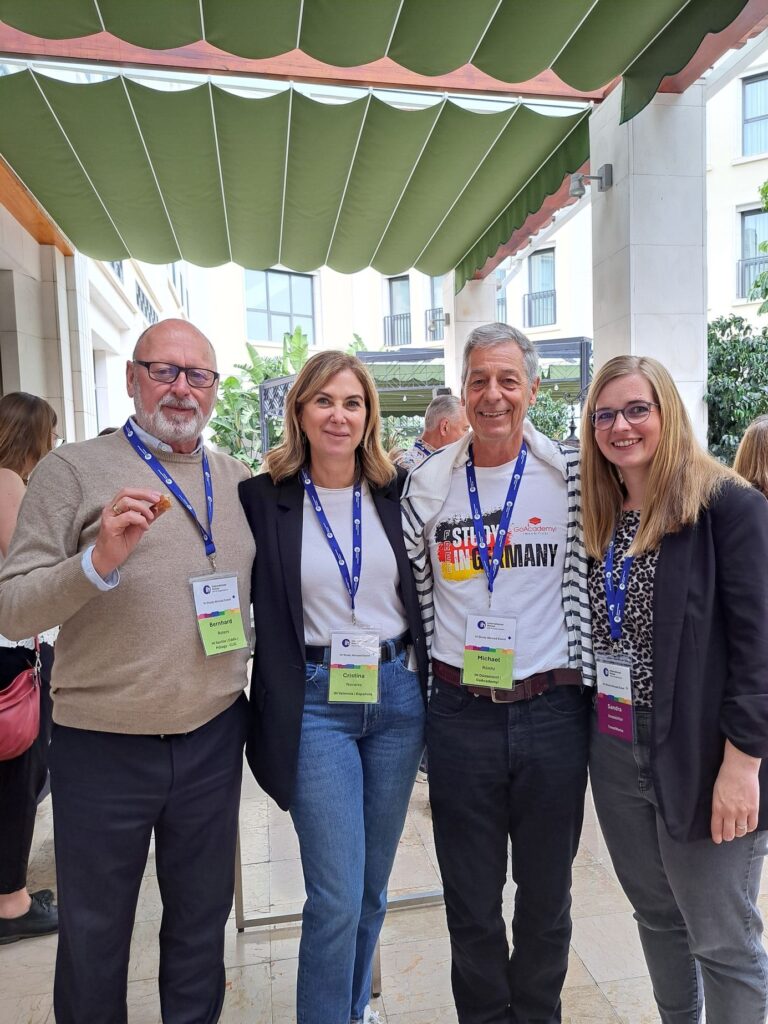 Four people wearing conference badges stand together indoors, smiling at the camera under a green striped awning.