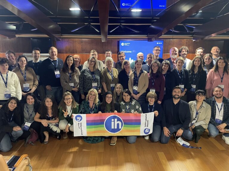 Large group of people posing indoors, some holding an IH International House banner with a rainbow design; conference name tags are visible on several attendees.