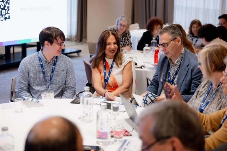 A group of people wearing name badges sit around a table engaged in discussion at a conference or workshop.