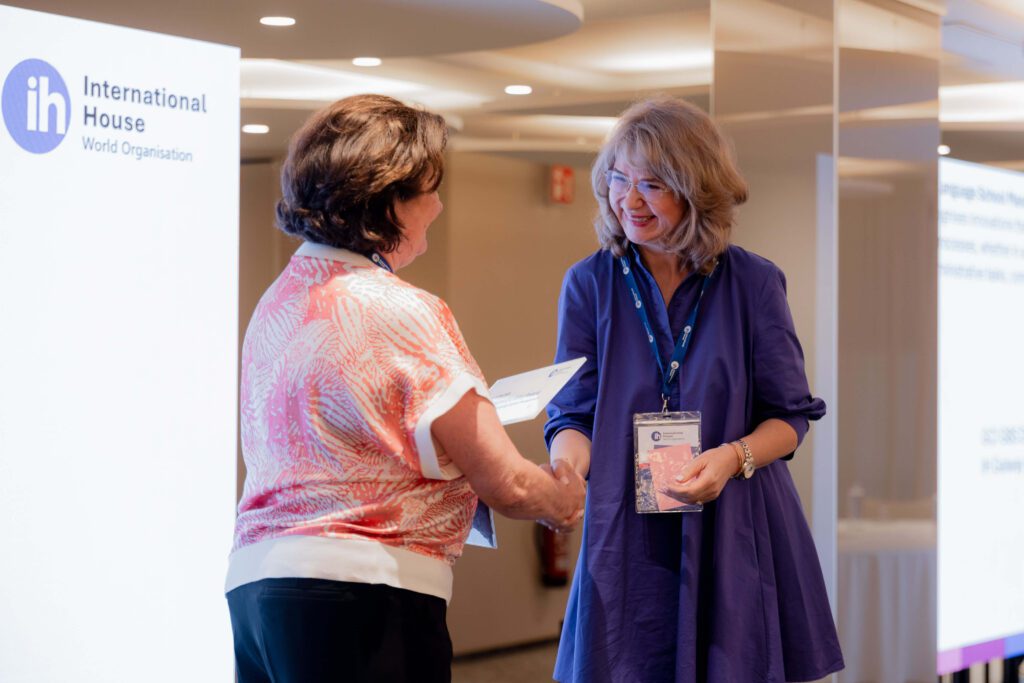 Two women shake hands in a conference setting next to an International House World Organisation sign, one holding a certificate and the other wearing a lanyard and badge.