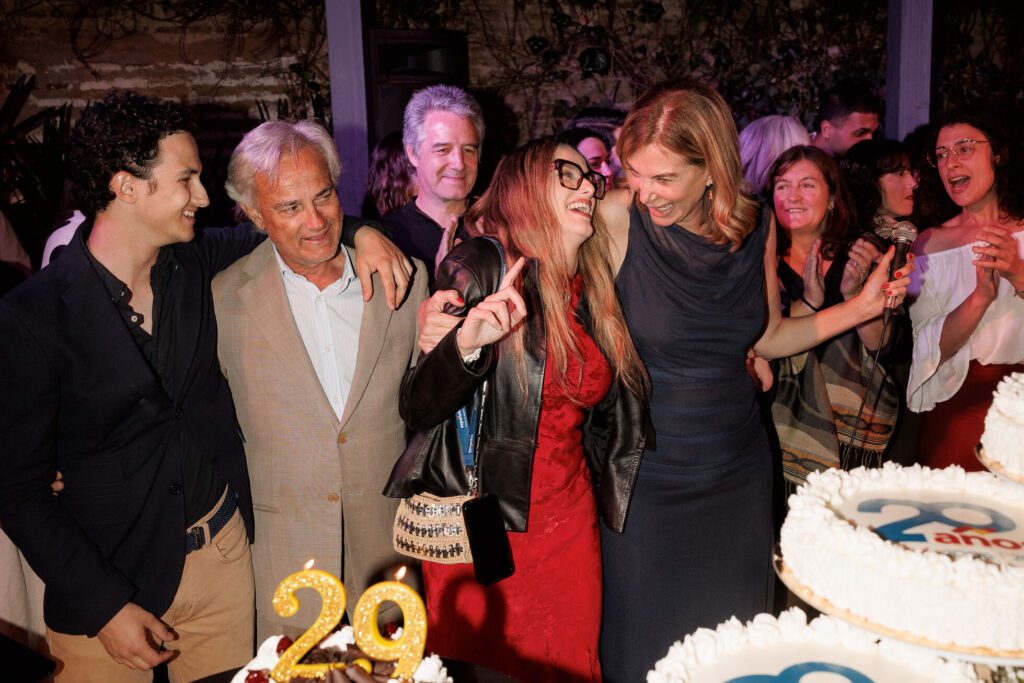 A group of people smiling and celebrating around cakes with "29" candles and "29 anos" decorations at a party.