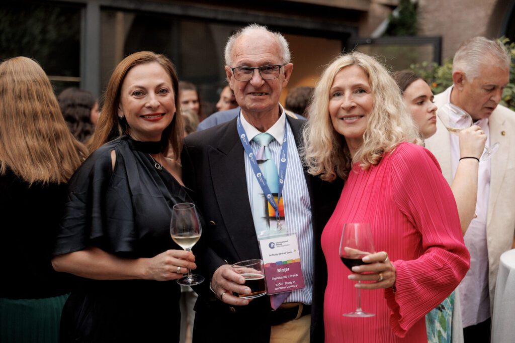 Three people stand together at an outdoor social event, each holding a drink. The man in the center wears a name badge and lanyards, while the two women smile at the camera.