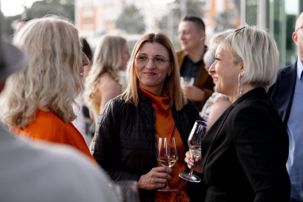 Three women stand together holding wine glasses and conversing at an outdoor social event, with other people visible in the background.