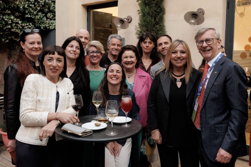 A group of people, dressed in business or semi-formal attire, pose and smile around a tall table with drinks and snacks at an indoor-outdoor event.