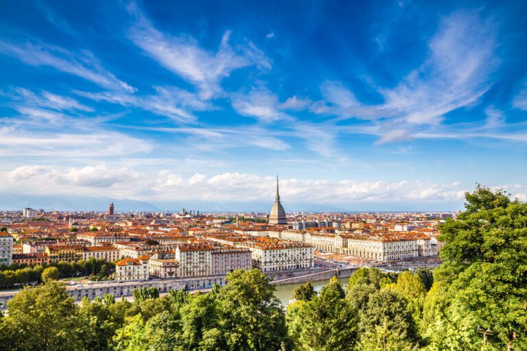 Panoramic view of Turin, Italy, showing the Mole Antonelliana, rows of buildings with red roofs, and the Po river under a blue sky with scattered clouds.