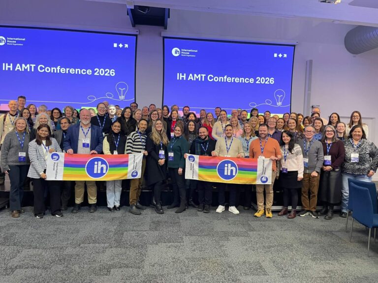 A large group of people pose indoors at the IH AMT Conference 2026, holding IH banners in front of two projection screens with the event name.