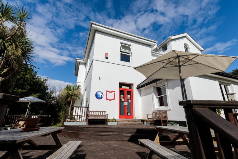 A white two-story building with a red door, outdoor seating, umbrellas, and blue sky with scattered clouds.