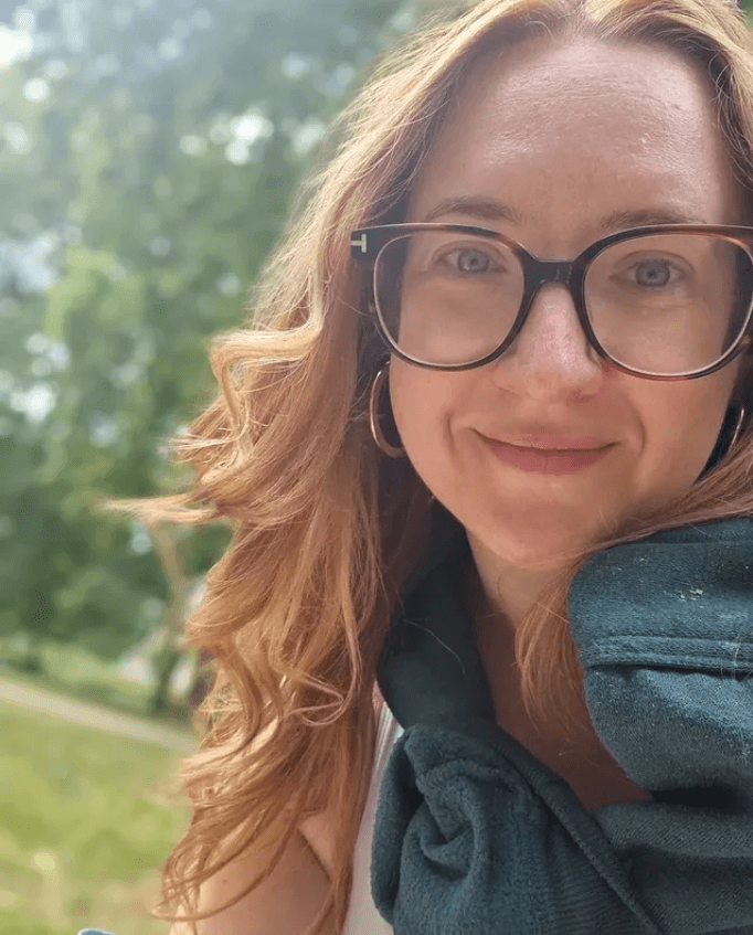 A woman with long red hair and glasses smiles outdoors, wearing a teal scarf. Green trees and a cloudy sky are visible in the background.