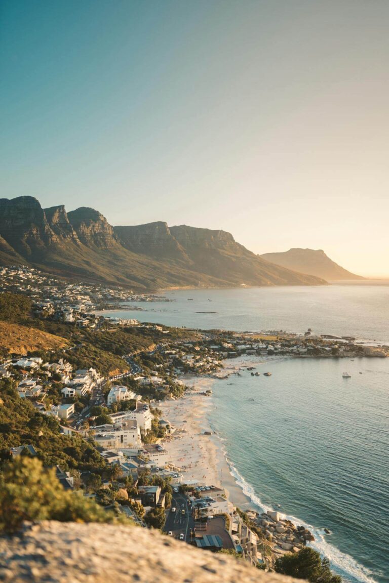 A coastal town with white sandy beaches and blue water sits at the base of green mountains under a clear sky at sunset.