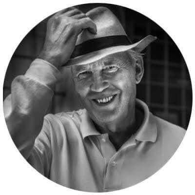 Older man in a light-colored shirt and hat smiling, tilting his hat with one hand. The photo is black and white and framed in a circular crop.