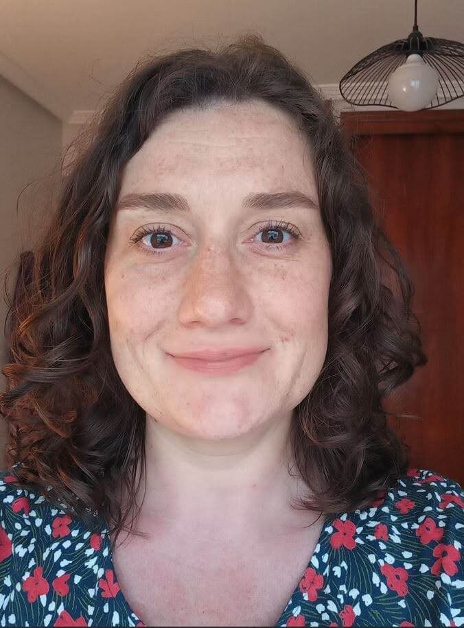 Woman with curly brown hair and floral top smiles at the camera indoors, with a wooden door and light fixture visible in the background.