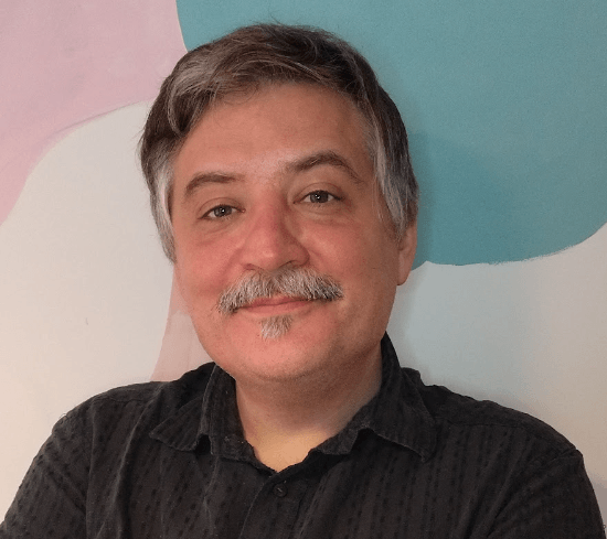 A person with short gray hair and a mustache is smiling at the camera, wearing a dark shirt, standing in front of a pastel-colored abstract background.