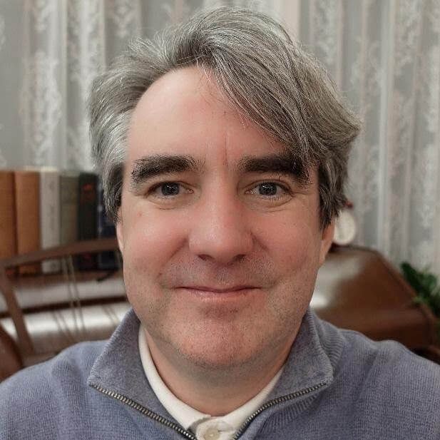 A middle-aged man with gray hair and a light beard smiles at the camera indoors; books and furniture are visible in the background.