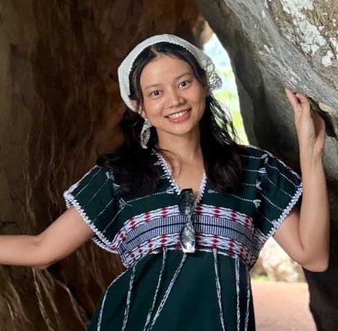 Woman standing in front of a rocky cave entrance, smiling, wearing a green patterned dress, white headscarf, and sunglasses hanging from her dress.