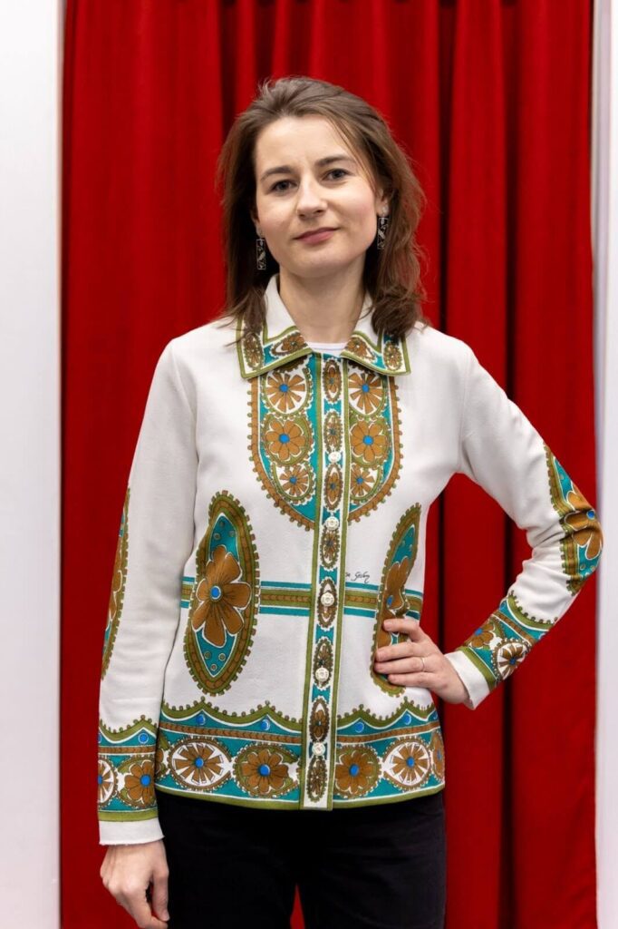 A woman stands in front of a red curtain, wearing a white shirt with green and gold geometric patterns, and black pants, with one hand on her hip.