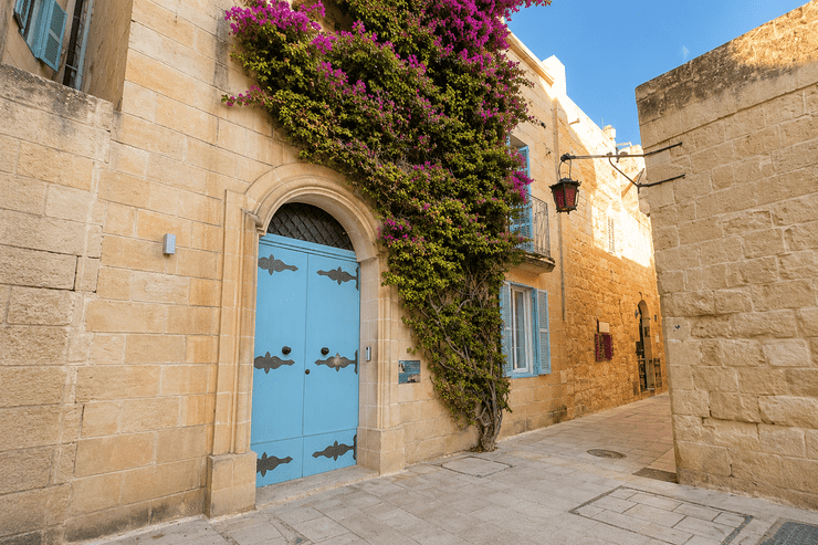 A narrow stone street flanked by traditional buildings, featuring a bright blue wooden door and vibrant purple bougainvillea climbing up the wall.