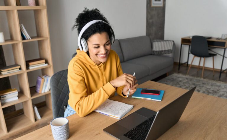 Woman in yellow hoodie wearing headphones, smiling while looking at a laptop. She is seated at a desk with notebooks, a phone, and a coffee mug in a living room setting.